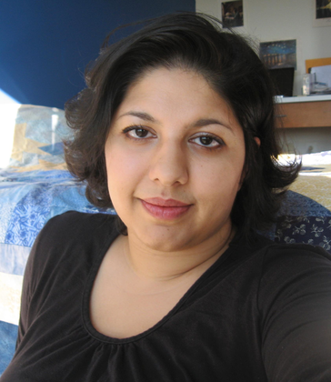 An Indian-American woman with short dark hair in a black top smiles at the camera, sitting in front of a homemade “night sky” quilt in her home office.