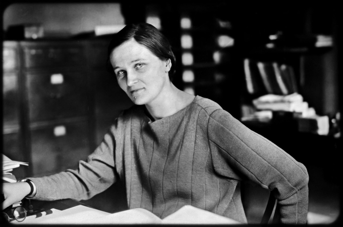 Black and white image of a caucasian British woman with short dark hair sitting in an office, looking at the camera, with one hand on her desk and one hand hooked around the back of her chair. On her desk are papers, an open book, and a pair of glasses.