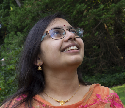An Indian woman with long dark hair, wearing glasses and a flowery pink top, smiles up towards the upper right of the photo in front of a background of leaves.