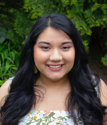 A Mexican-American woman with long black hair in a white and green flowery top smiles at the camera in front of a background of leaves.